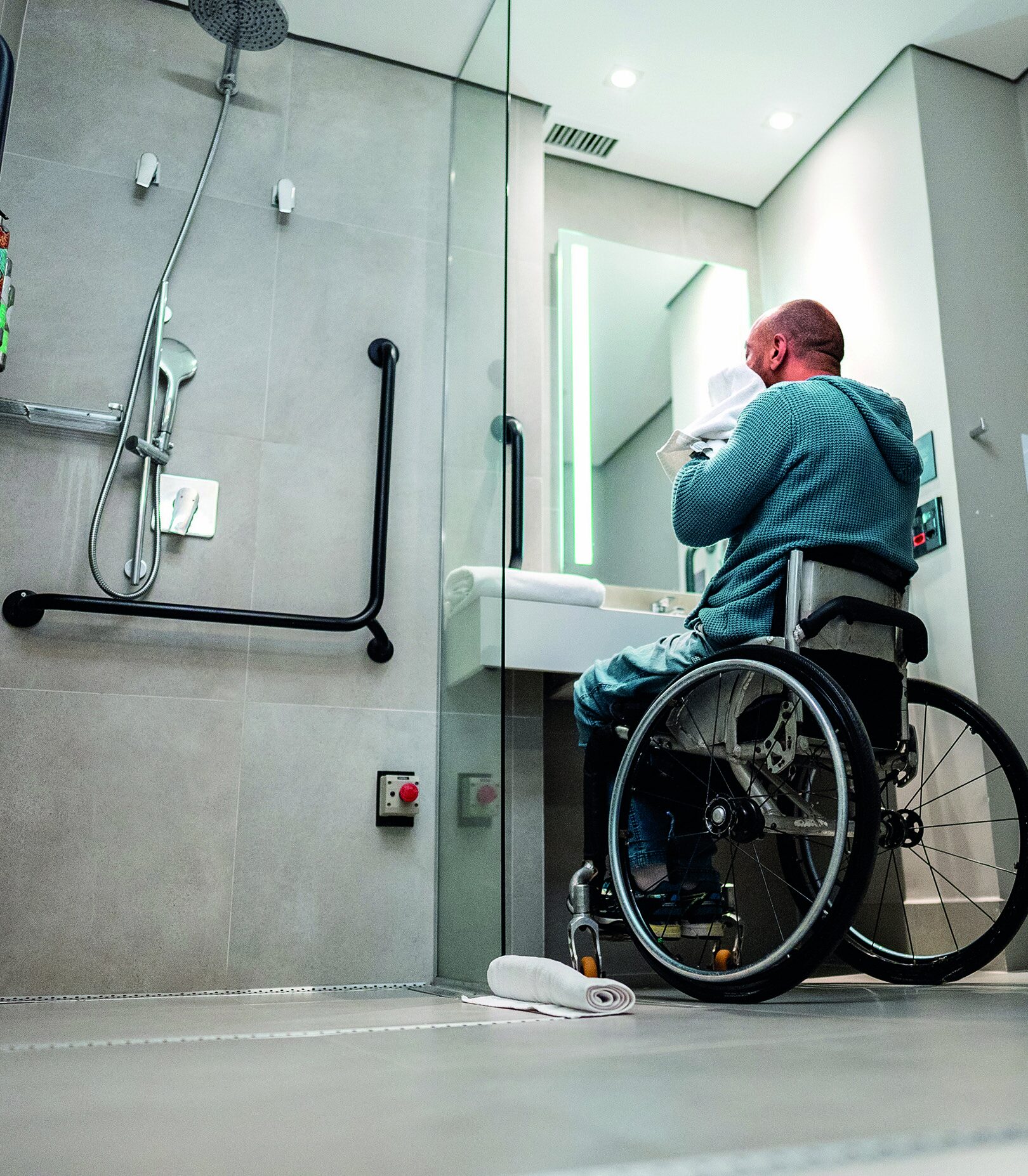 A person using a wheelchair washes their face at a sink in an adapted bathroom