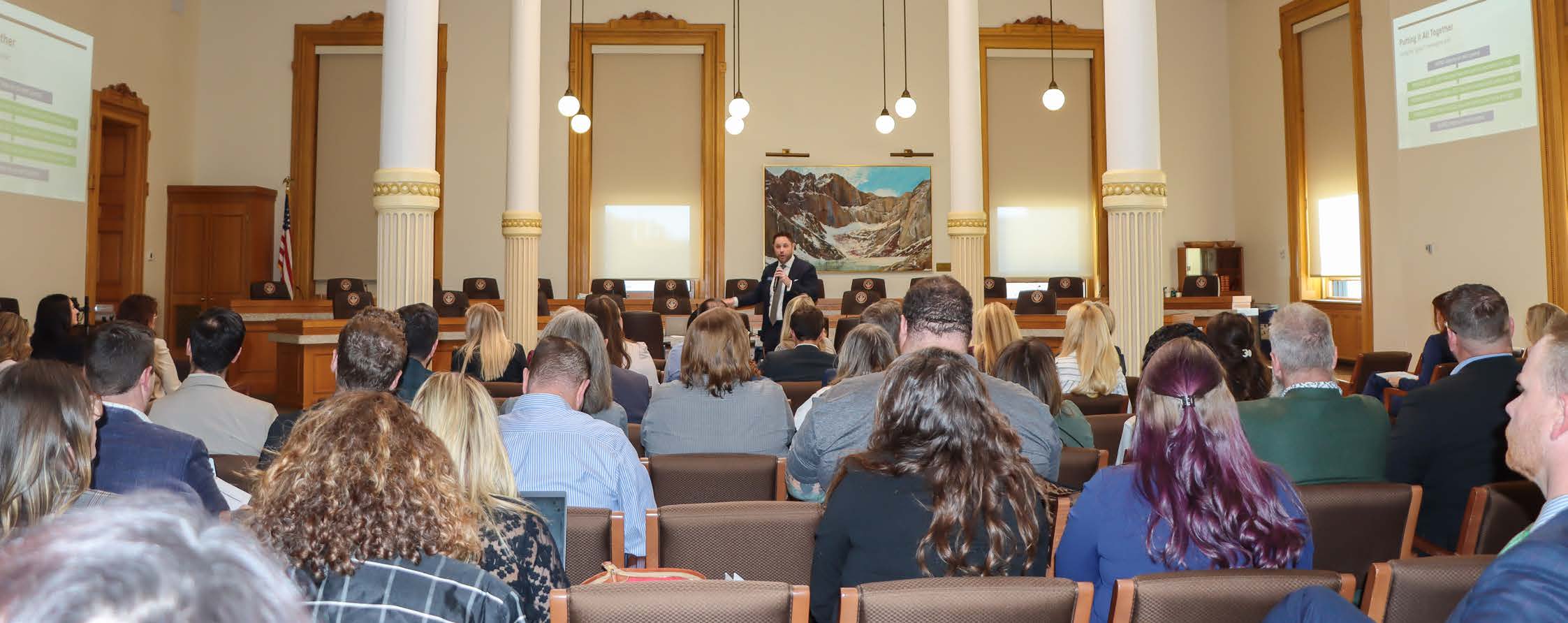 A group of industry leaders gathered in a grand, historic meeting room at the Capitol, listening to a presentation.